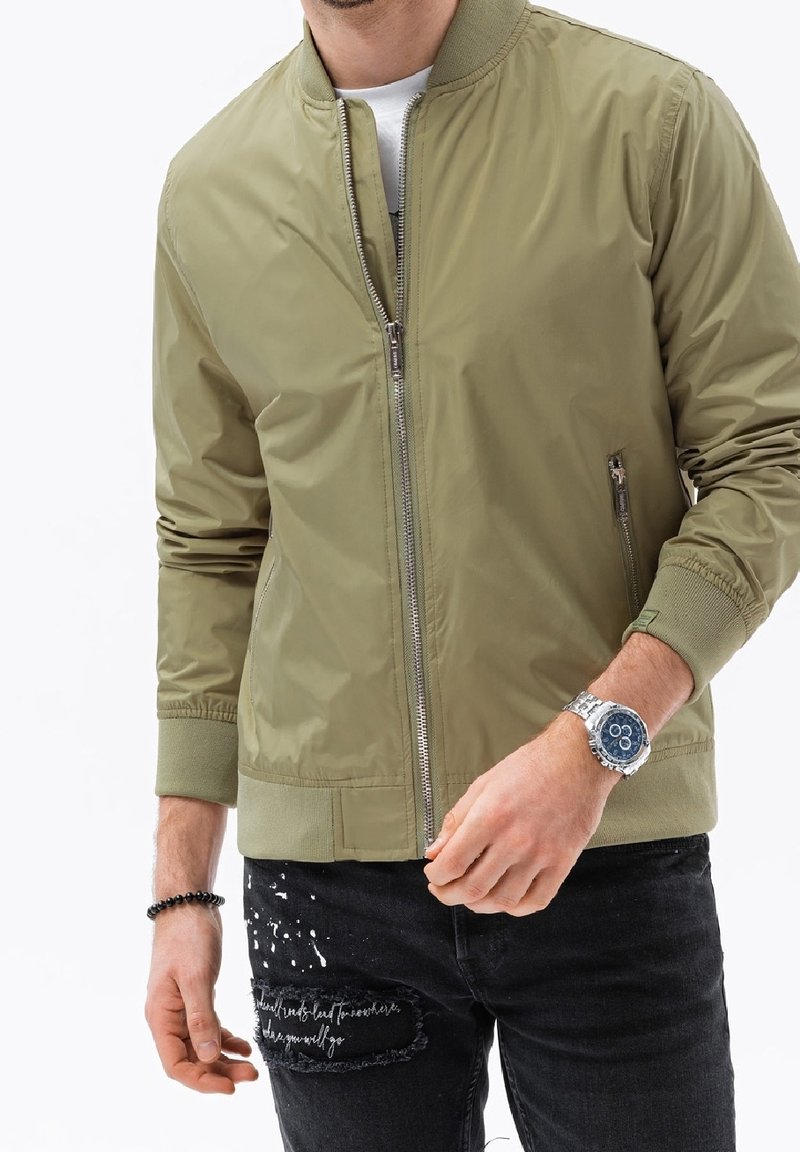 Homme portant une veste bomber vert olive, un jean noir avec des éclaboussures de peinture blanche et un patch, une montre argentée et un bracelet de perles noires.