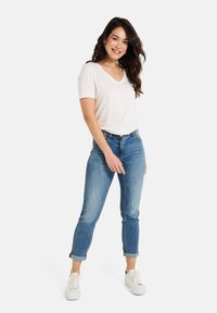 T-shirt blanc en V porté avec un jean ajusté bleu clair, à manches retroussées, et des baskets blanches. Texture de tissu lisse, style décontracté.