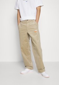 Beiga kordflorsbyxor med rak passform och synliga fickor, prydda med en orange Nike-logotyp på vänster sida. Kombinerade med vita sneakers.