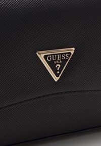Must mustrakk nahk käekott, millel on kuldne kolmnurkne logo, millel on kiri "GUESS USA" ja küsimärk. Lame siluett koos minimaalsete aktsentidega.