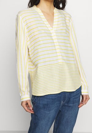 Woman wearing a white long-sleeve blouse with yellow stripes and blue jeans, standing with hands relaxed by sides.