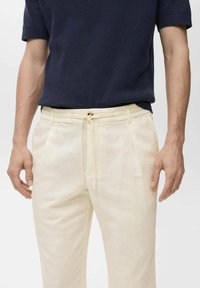 Pantalones de lino beige claro con cintura elástica y cordón. Presentan bolsillos laterales, corte relajado y una tela suave y texturizada.
