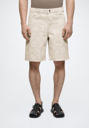 Mann iført beige blomstrete knelengde shorts og svarte åpne skinnsandaler, står mot en ensfarget bakgrunn.