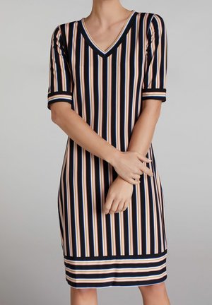 Kvinde iført knælange kjole med korte ærmer og lodrette striber i sort, hvid, orange og blå samt V-hals, hænderne foldet foran.