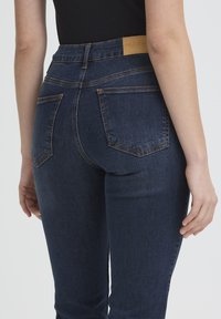 Gros plan sur une personne portant un jean taille haute bleu foncé avec des poches arrière et un haut noir, debout, les bras détendus le long du corps.