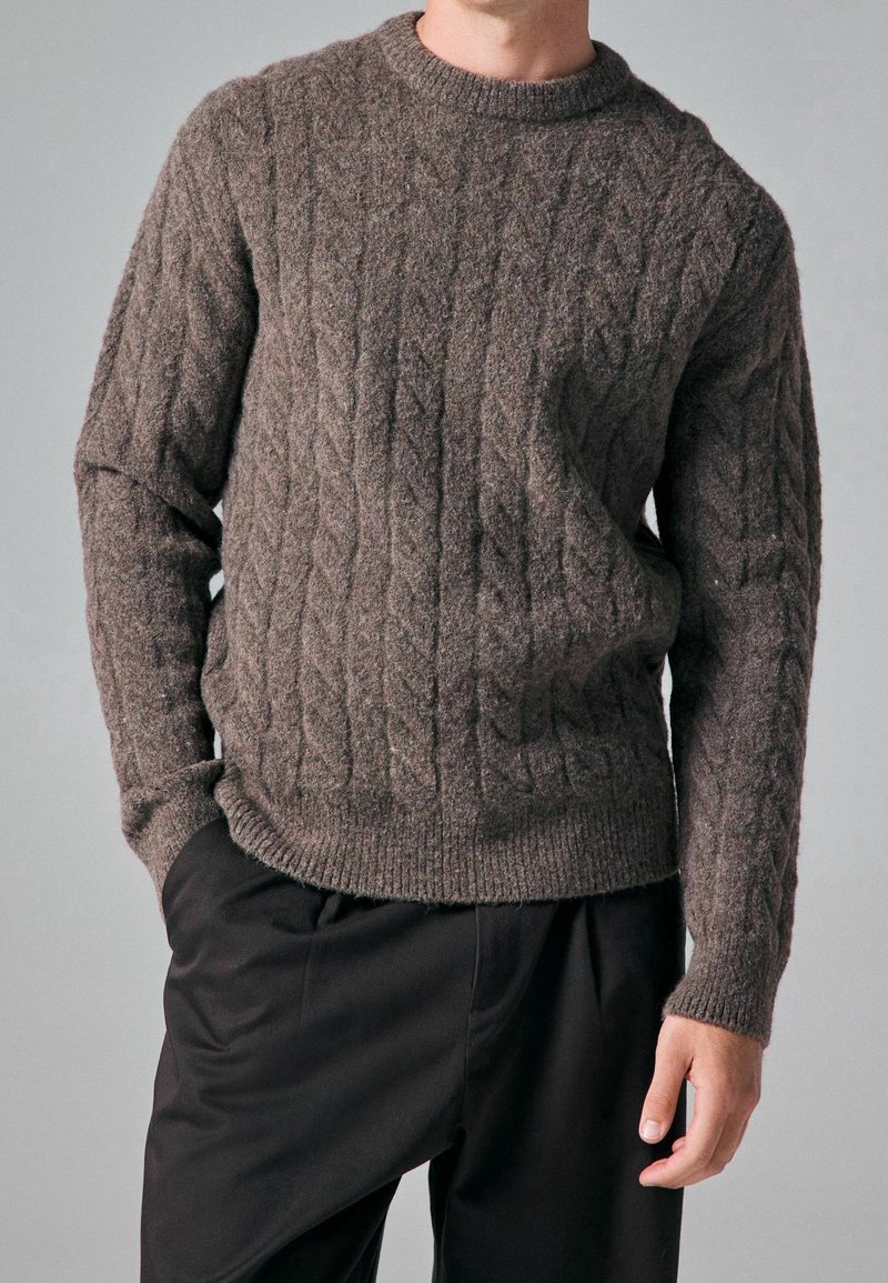 Maglione marrone a maglia a trecciola con scollatura rotonda, caratterizzato da motivi testurizzati e orlo a costine; indossato sopra pantaloni neri.