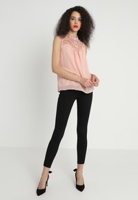 Ermeløs rosa bluse med blondebesetning ved halsen og kanten, kombinert med svarte skinny bukser og svarte høye hæler.