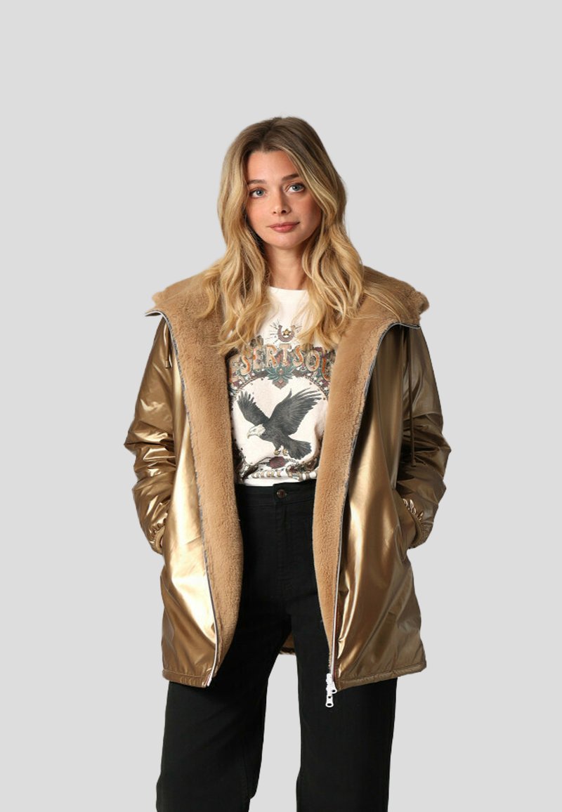 Parka doré métallisé avec un col en fausse fourrure, fermeture éclair et poches latérales. Portée sur un t-shirt graphique et un pantalon noir.