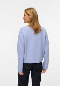 Gaiši zils adīts džemperis ar brīvu piegriezumu, platas piedurknes, ribota apakšmala un vienkārša apaļa kakla izgriezums, valkāts kopā ar tumšām džinsa biksēm.