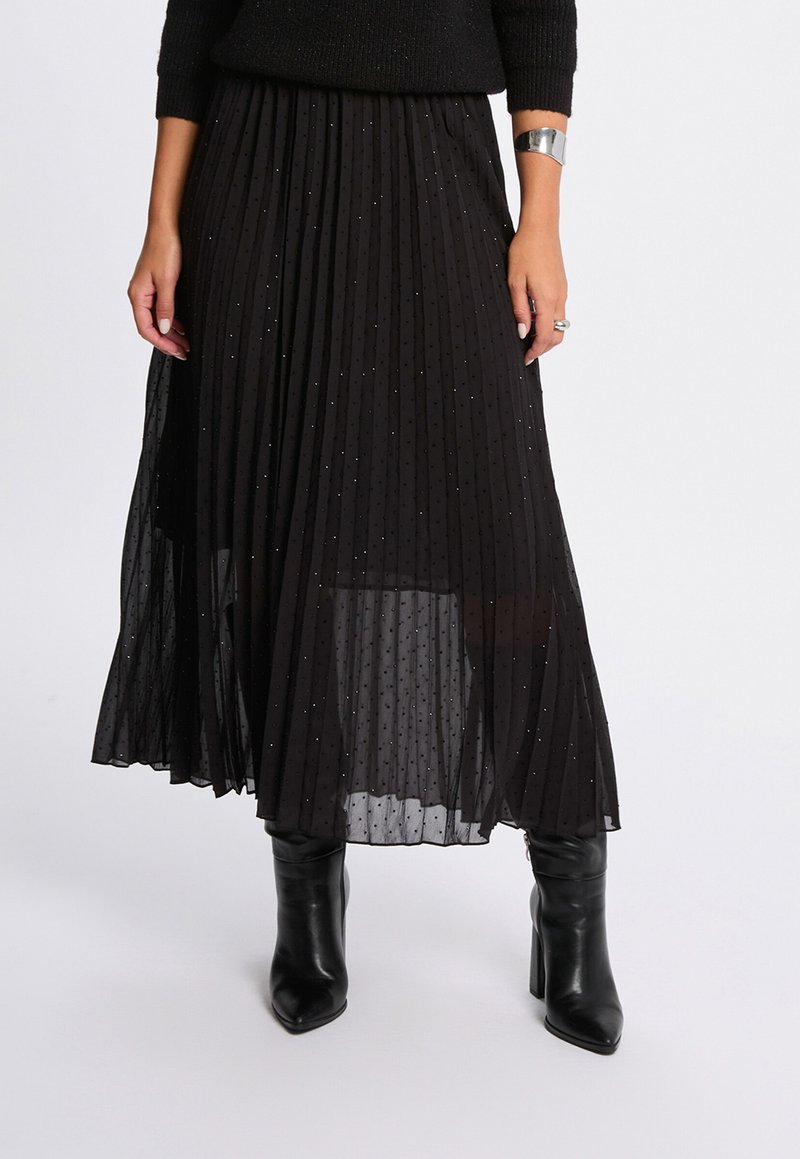 Sort plisseret nederdel med et gennemsigtigt overlay, prydet med små glimmer, lagdelt for tekstur, parret med sorte hælede støvler.
