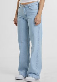 Lys blå vide bukser laget av denim, med høy midje, frontlommer og synlig søm. Brukt med hvite sneakers.
