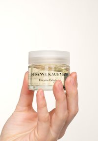 Przezroczysty szklany słoik z półprzezroczystą białą pokrywką, zawierający żółty peeling enzymatyczny. Na słoiku znajduje się tekst "SUSANNE KAUFMANN" oraz "Enzyme Exfoliator."