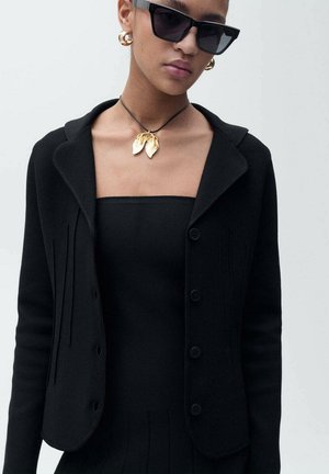 Black knitted cardigan with a collar and button closure, paired with a square neck top. Features gold leaf pendant on a black cord necklace.