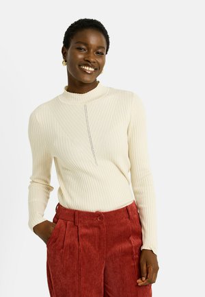 Pull tricot côtelé crème avec un col montant et un détail de couture à l'avant, associé à un pantalon en velours côtelé rouge texturé avec une fermeture à boutons.