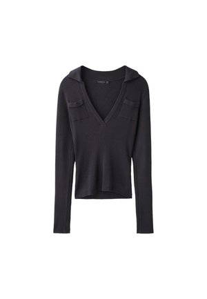 Femme portant une veste en cuir grise, un haut noir sans manches, un large pantalon culotte noir et des talons noirs pointus, debout devant un fond gris.