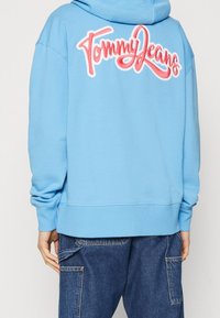 Ljusblå hoodie med en stor broderad "Tommy Jeans"-logotyp i rosa på ryggen. Har en känguruficka och ribbade ärmslut.