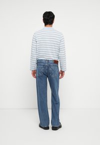 Gestreiftes Langarmhemd in Hellblau und Weiß; lockere blaue Jeans mit zwei Gesäßtaschen und einem braunen Lederpatch am Bund.