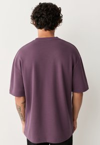 Lila Baumwoll-T-Shirt mit kurzen Ärmeln und entspannter Passform. Glatte Textur und rundem Halsausschnitt. Sichtbares Tattoo am Unterarm.