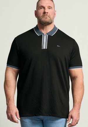Skægget mand iført en sort kortærmet polo med en mørk krave med hvide striber og lyseblå jeans, stående.
