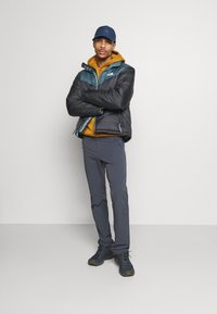 Giubbotto imbottito nero e verde acqua, felpa gialla, pantaloni grigi, cappellino blu navy e sneaker scure, tutti con varie texture e materiali.