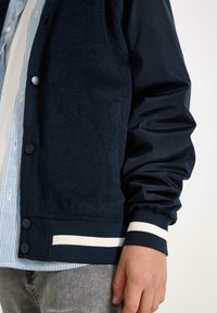 Marineblaue Varsity-Jacke mit Wollkörper und glänzenden Nylonärmeln. Ausgestattet mit Druckknöpfen und gestreiften Rippbündchen sowie Saum.