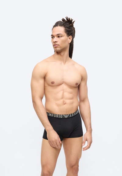 Άνδρας χωρίς πουκάμισο με dreadlocks, φορώντας μαύρα boxer briefs Emporio Armani, στέκεται μπροστά σε απλό λευκό φόντο, κοιτάζοντας προς το πλάι.