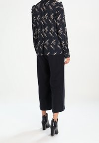 Žena v černé květinové blůze, širokých černých kalhotách po kotníky a černých podpatkových kotníkových botách, stojící zády k bílé pozadí.