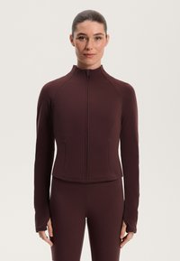 Burgundi színű, zippes dzseki, testhez simuló fazonnal, hosszú ujjakra és hüvelykujjlyukakkal. Simító, elasztikus anyagból készült, ideális aktív viselethez.