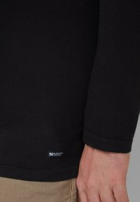 Zwart shirt met lange mouwen en geribbelde textuur, met een klein blauw label met de tekst "Elevated Casual" nabij de zoom. Subtiele stikdetail.