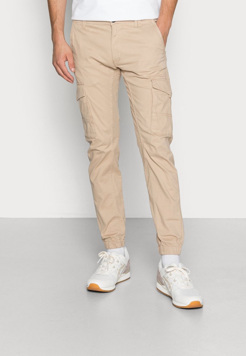 Man in beige cargobroek met elastische manchetten, witte sneakers en witte sokken, staand met handen in de zakken op een grijze vloer.