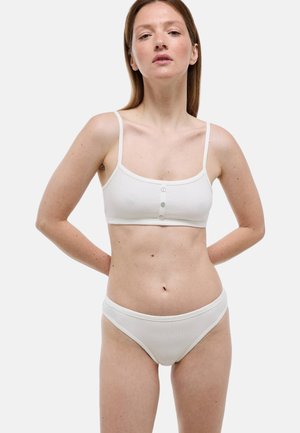 Femme portant un bralette côtelé blanc avec des boutons et un sous-vêtement assorti côtelé blanc, debout les mains derrière le dos sur un fond clair.