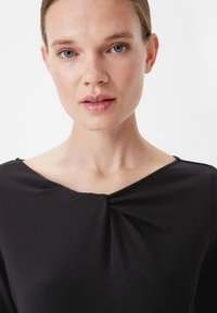 Schwarzes Oberteil mit V-Ausschnitt und Raffung in der Mitte. Glattes Material, lange Ärmel und eine figurbetonte Silhouette. Sauberes, minimalistisches Design.