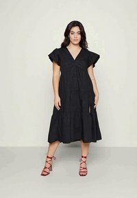 Mulher a ficar de pé contra um fundo simples, vestindo um vestido midi preto com decote em V e mangas godê, combinado com sandálias vermelhas de tiras.