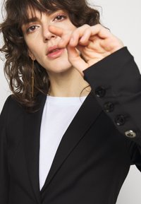 Blazer noir ajusté avec un col prononcé, un tissu texturé, des poignets boutonnés, porté par-dessus un T-shirt blanc col rond.