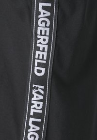 Melns audums ar baltu logo joslu, kurā ir rakstīts "LAGERFELD" un "by KARL LAGERFELD" gar sānu. Gluda tekstūra un sportisks stils.