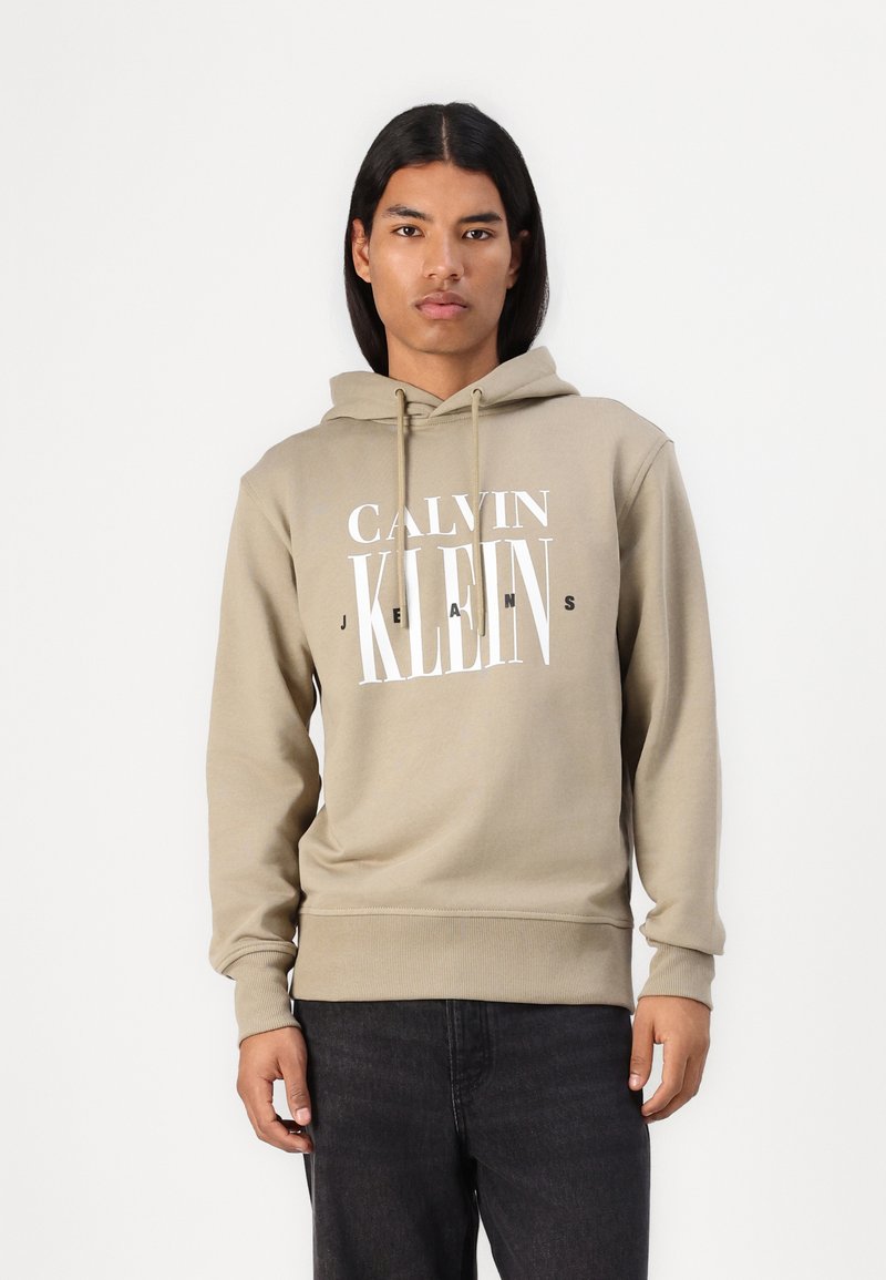 Sweatshirt cor caqui feito de mistura de algodão, apresentando uma graphic frontal "CALVIN KLEIN JEANS" em branco e preto, com capuz ajustável por cordão e punhos em canelado.