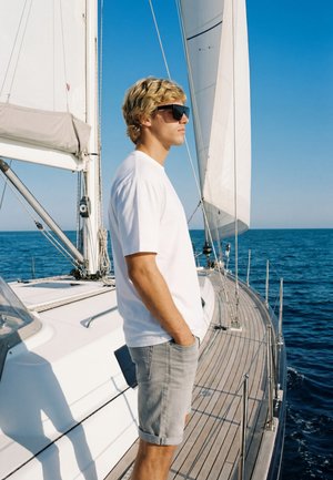 Jeune homme portant des lunettes de soleil et des vêtements décontractés, debout sur le pont d'un voilier, regardant la mer bleue calme sous un ciel clair.