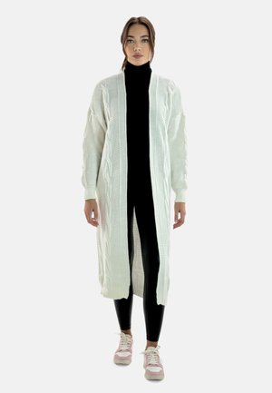 Lang cardigan med åben front i ribbet creme strik med kabeldetaljer. Bæres over en sort turtleneck og sorte leggings, matchet med lyserøde sneakers.