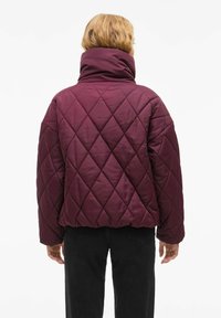 Bordo jakna sa duksericom, s visokim ovratnikom, s dijamantnim uzorcima i elastičnim manžetama, u kombinaciji s crnim hlačama.