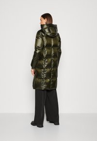 Dlhý, vypchávkovaný kabát s lesklou tmavozelenou farbou a kapucňou, s prešívaným dizajnom a bočnými zipsami. Čierne nohavice dotvárajú outfit.