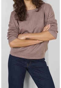 Maglione a coste di un marrone chiaro con maniche ampie, indossato con jeans blu scuro. Il maglione presenta un collo rotondo e una vestibilità comoda.