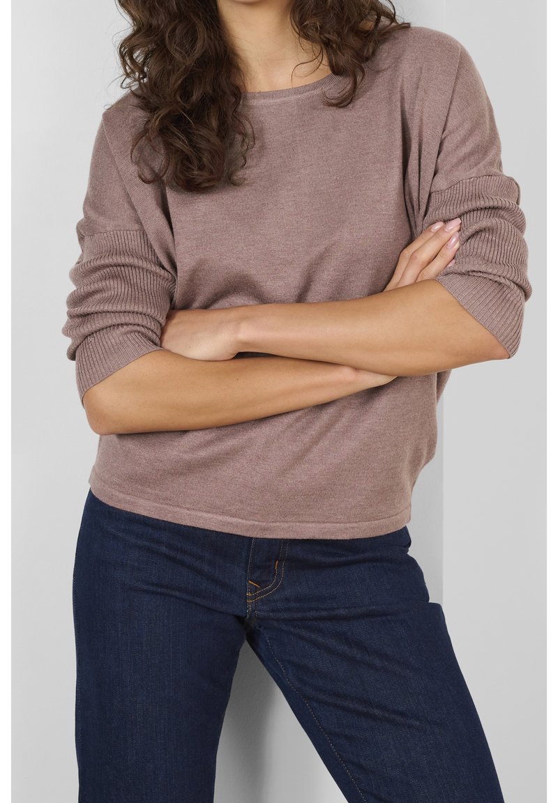 Maglione a coste di un marrone chiaro con maniche ampie, indossato con jeans blu scuro. Il maglione presenta un collo rotondo e una vestibilità comoda.