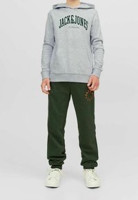Felpa grigia con "JACK&JONES" in testo verde, abbinata a pantaloni della tuta verde scuro con un logo arancione. Sneakers bianche completano l'outfit.