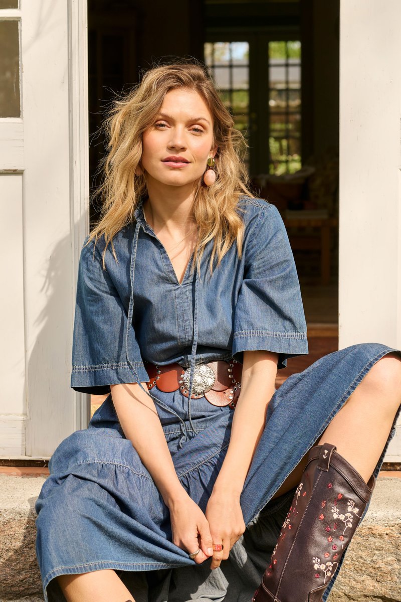 Femme blonde en robe en denim avec une large ceinture assise sur des marches, portant des bottes marron brodées de fleurs, semblant détendue au soleil.