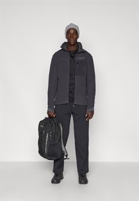 Giacca in pile grigia con dettagli neri, tasca sul petto con zip, pantaloni neri, berretto grigio e zaino nero con cerniere multicolori.