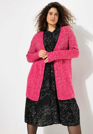 Cárdigan de punto rosa con cierre frontal y mangas largas, combinado con un vestido negro que presenta un sutil patrón de leopardo. Textura suave y ajuste relajado.