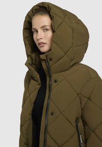 Gequilted olijfgroene pufferjack met een grote capuchon, ritssluiting aan de voorkant en knoopsluiting, met zwarte accenten en twee zijzakken.