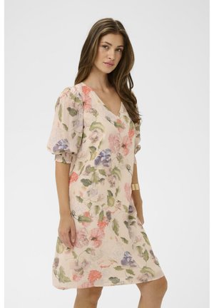 Kvinde iført en lyserød kjole med puffede ærmer og flerfarvet blomsterprint, stående mod en ensfarvet hvid baggrund.