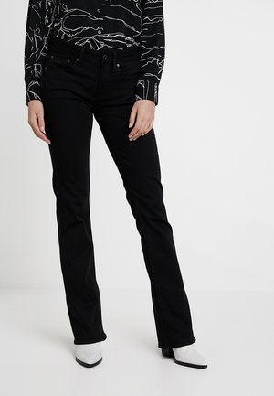 Femme portant un jean noir ajusté, une chemise noire à manches longues à motifs et des bottes blanches à bout pointu, debout sur un fond blanc.