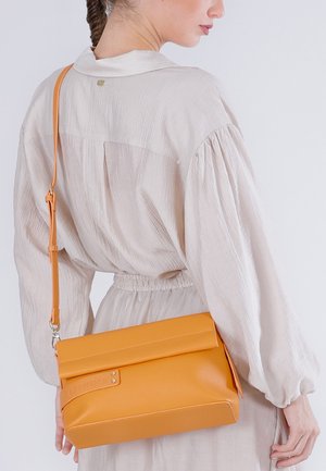 Vrouw met gevlochten haar en een lichtbeige jurk draagt een middelgrote oranje leren schoudertas op haar rug.